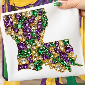 May include: A white square item featuring a jeweled outline of the state of Louisiana. The outline is filled with green, purple, and clear gemstones, with gold-colored accents. The item is held by a person with green nail polish.