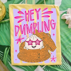 May include: Yellow tote bag with the text "HEY DUMPLING" in pink. The design features a cartoon dumpling wearing star-shaped sunglasses, inside a bamboo steamer basket. Pink stars and stripes complete the design.