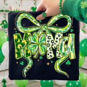 Pode incluir: Camiseta preta com um laço verde e amarelo e a palavra "MAMA" em um design de trevo. As letras apresentam uma variedade de padrões, incluindo floral e estampa de leopardo. A camiseta é segurada por uma pessoa usando uma pulseira e um anel verdes.