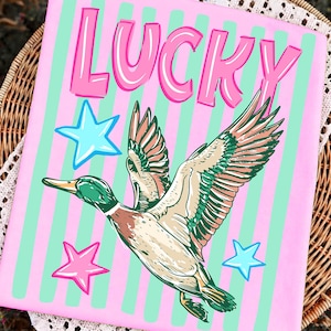 Puede incluir: Fondo rosa a rayas verdes con la palabra &laquo;LUCKY&raquo; en rosa. Ilustraci&oacute;n de un pato volador con plumas verdes, marrones y blancas, adem&aacute;s de estrellas azules y rosas.