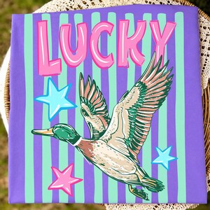 Puede incluir: Camiseta morada con rayas verticales en verde menta y morado. La palabra "LUCKY" est&aacute; en letras rosas con burbujas. Una ilustraci&oacute;n de un pato volador est&aacute; en el centro, con estrellas rosas y azules.