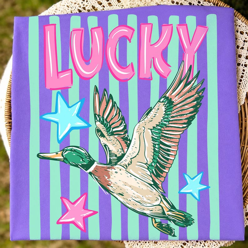 Puede incluir: Camiseta morada con rayas verticales en verde menta y morado. La palabra "LUCKY" est&aacute; en letras rosas con burbujas. Una ilustraci&oacute;n de un pato volador est&aacute; en el centro, con estrellas rosas y azules.