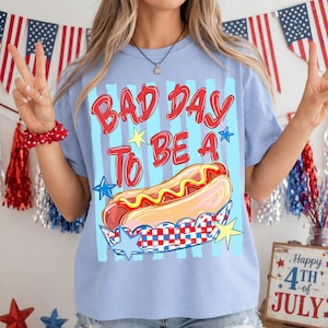以下が含まれることがあります： 赤色の「BAD DAY TO BE A」の文字とマスタードを添えたホットドッグのグラフィックが特徴の、水色のTシャツ。 ホットドッグは赤、白、青のチェック柄の紙トレイに入っています。 背景には星とストライプがあります。
