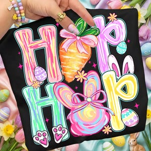 Peut inclure: T-shirt noir avec les mots "Hip Hop" en lettres color&eacute;es. Le motif comprend une carotte, des oreilles de lapin, un lapin et des &oelig;ufs de P&acirc;ques. Le design est aux couleurs vives avec des accents roses, jaunes et bleus.