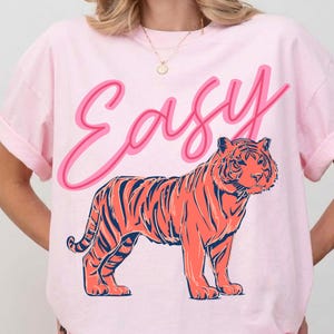 May include: A light pink t-shirt featuring a stylized orange tiger with navy blue stripes. The word "Easy" is written above the tiger in a pink script font. The shirt has rolled-up sleeves and a crew neck.