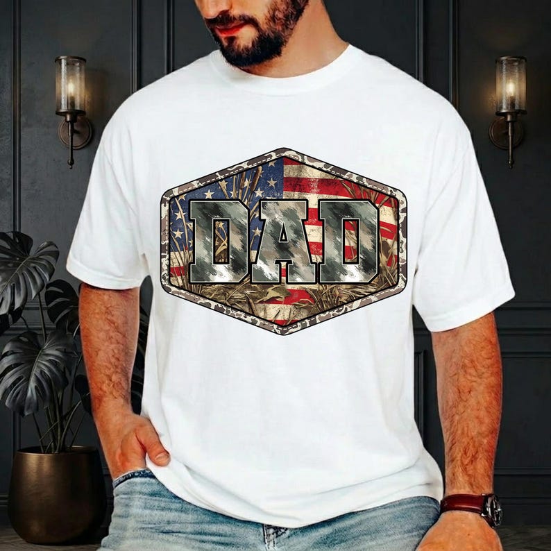 以下が含まれることがあります： 白いTシャツ。六角形のグラフィックデザイン。グラフィックは、迷彩とアメリカ国旗のデザインを背景に、テクスチャードフォントで「DAD」という単語を表示します。カジュアルウェアアイテム。