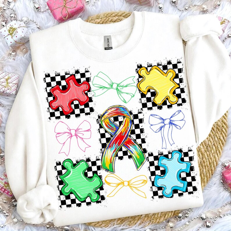 May include: White sweatshirt with a design of colourful puzzle pieces and bows on a black and white chequered background. A rainbow ribbon is also featured. The design includes red, green, yellow, pink, blue, and orange elements. The sweatshirt has a crew neck and long sleeves.