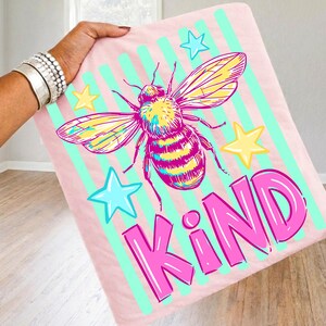 Puede incluir: Manta rosa con una gran ilustraci&oacute;n de abeja, estrellas y la palabra "KIND" en rosa brillante. La abeja tiene detalles amarillos y morados, sobre un fondo de rayas verticales verde claro. Un dise&ntilde;o divertido y colorido.