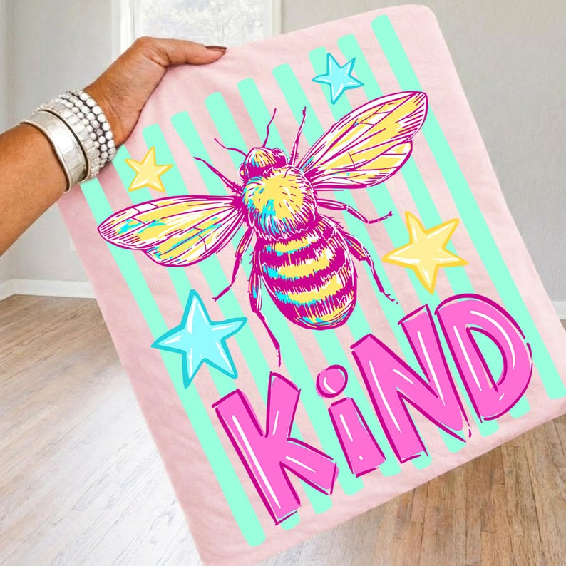 Puede incluir: Manta rosa con una gran ilustraci&oacute;n de abeja, estrellas y la palabra "KIND" en rosa brillante. La abeja tiene detalles amarillos y morados, sobre un fondo de rayas verticales verde claro. Un dise&ntilde;o divertido y colorido.
