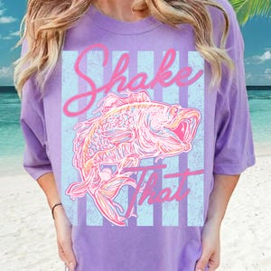 K&ouml;nnte beinhalten: Ein lila T-Shirt mit einer Fischgrafik und dem Schriftzug "Shake That" in rosa Schrift. Der Fisch ist mit rosa, orangefarbenen und blauen Linien detailliert, vor einem hellblau-wei&szlig; gestreiften Hintergrund.