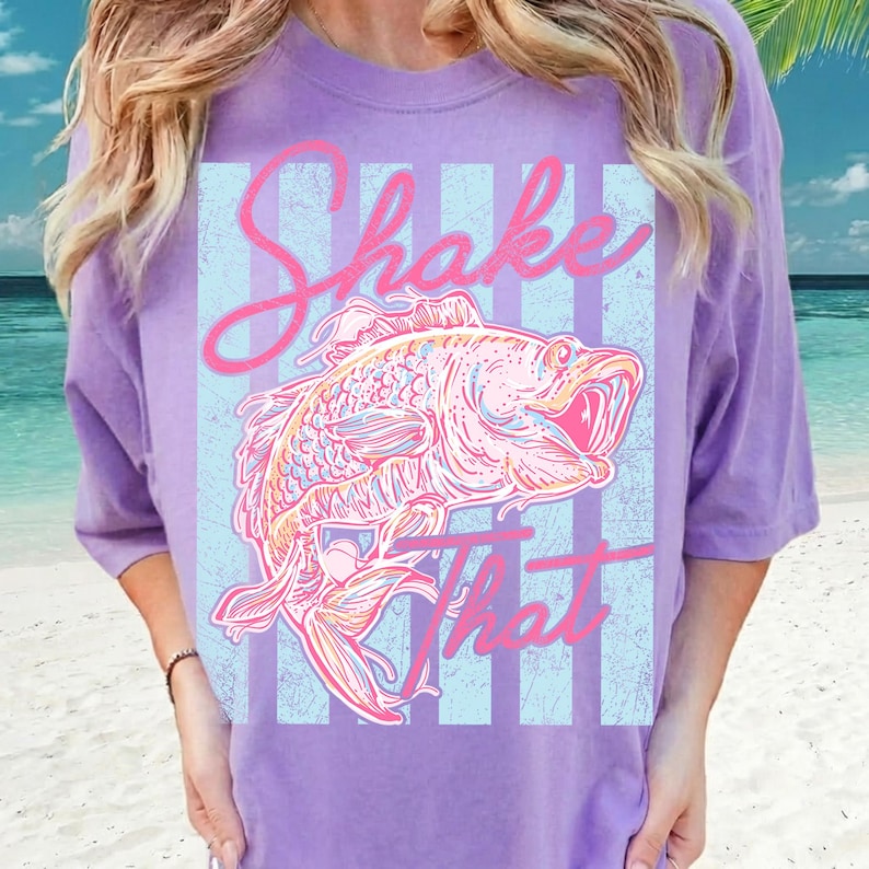 K&ouml;nnte beinhalten: Ein lila T-Shirt mit einer Fischgrafik und dem Schriftzug "Shake That" in rosa Schrift. Der Fisch ist mit rosa, orangefarbenen und blauen Linien detailliert, vor einem hellblau-wei&szlig; gestreiften Hintergrund.