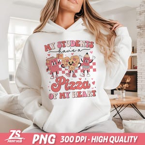 K&ouml;nnte beinhalten: Wei&szlig;er Hoodie mit einem rosafarbenen Grafikdesign, auf dem "Meine Sch&uuml;ler haben eine Pizza meines Herzens" steht. Die Grafik zeigt ein Buch, ein Pizzast&uuml;ck und einen Bleistift mit Herzen.