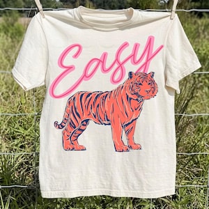 May include: Cream-colored t-shirt with the word "Easy" in pink script above a coral-colored tiger with navy blue stripes. The t-shirt is hanging on a clothesline.