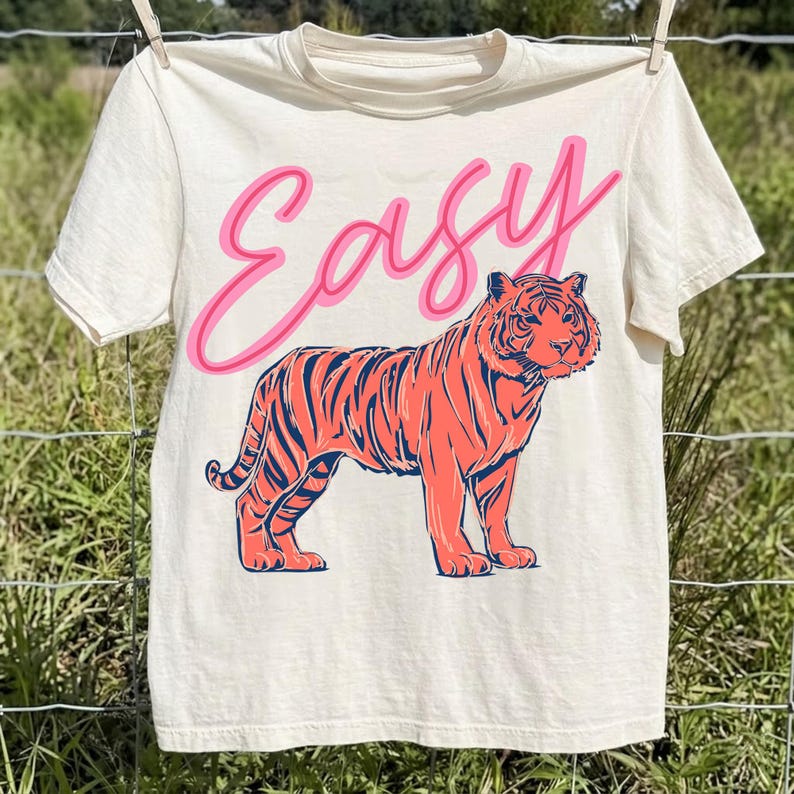 May include: Cream-colored t-shirt with the word "Easy" in pink script above a coral-colored tiger with navy blue stripes. The t-shirt is hanging on a clothesline.