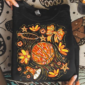 May include: Black sweatshirt featuring a basketball design with a basketball, hoop, flowers, stars, and a bow. The basketball is orange with gold accents, and the flowers and stars are orange and yellow.