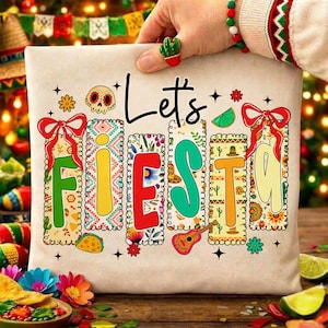 Puede incluir: Un cojín beige con la frase "Let's Fiesta" en escritura negra y letras de colores. Las letras están adornadas con imágenes festivas como una calavera, flores y una guitarra. Los lazos rojos y las estrellas realzan el tema festivo.