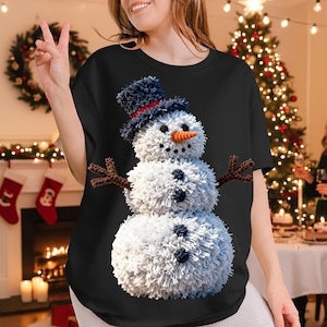 May include: Black t-shirt featuring a snowman design. The snowman has a blue hat with a red band, a carrot nose, and twig arms. The snowman is made of white, fluffy material with dark blue buttons. Christmas theme.