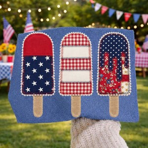 May include: A blue towel with three decorative popsicle designs. The popsicles feature red, white, and blue patterns, including stars, gingham, and floral prints. Each popsicle has a burlap stick and a scalloped edge.