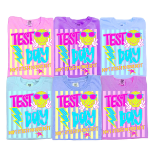 Op de afbeelding: Zes pastelkleurige t-shirts met de woorden "TEST DAY" in een gedurfd, kleurrijk ontwerp. Elk shirt heeft een lachend gezicht met een zonnebril, een bliksemschicht en de tekst "DON'T STRESS DO YOUR BEST".