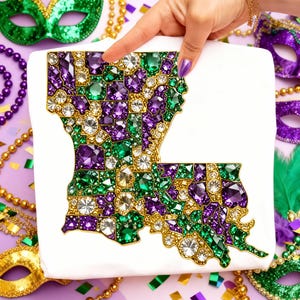 May include: A white square-shaped item featuring a jeweled outline of the state of Louisiana. The design is filled with purple, green, and clear gemstones. The item is surrounded by Mardi Gras beads and masks.