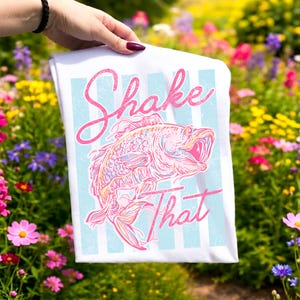 K&ouml;nnte beinhalten: Wei&szlig;es T-Shirt mit einem rosa und blauen Grafikdesign eines aus dem Wasser springenden Fisches. Der Text "Shake That" steht &uuml;ber und unter dem Fisch. Das Design hat einen Vintage-Look.