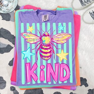 Puede incluir: Apilados camisetas en tonos morado, rosa y naranja. La camiseta superior es morada con un gr&aacute;fico de una abeja, estrellas y la palabra "KIND" en rosa. La abeja tiene alas amarillas y rosas. La camiseta tiene rayas verdes verticales.