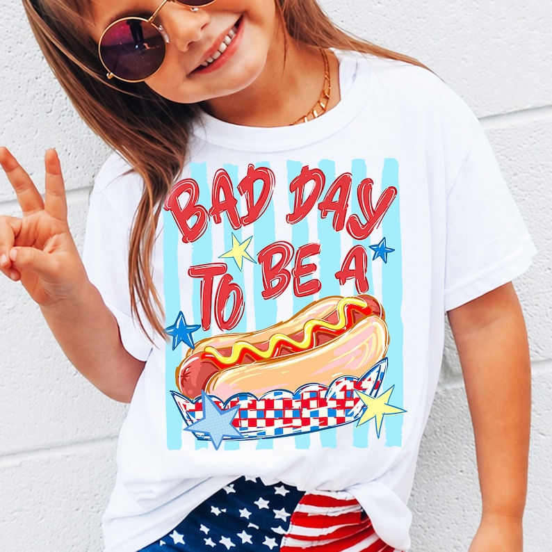 以下が含まれることがあります： 赤い文字で「BAD DAY TO BE A」と書かれた白いTシャツと、ホットドッグのイラスト。 ホットドッグは赤、白、青のチェック柄の容器に入っており、青と黄色の星のアクセントが付いています。 シャツには青い縦縞があります。