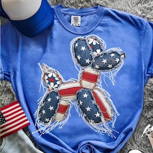 May include: Blue t-shirt featuring a balloon dog design in red, white, and blue, with star and floral patterns. The design is reminiscent of the American flag. A blue and white baseball cap and an American flag patch are also visible.