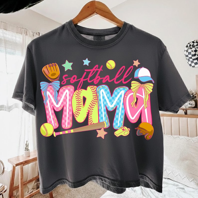 May include: Dark gray t-shirt with the words "softball MAMA" in colorful, playful lettering. The design includes a baseball glove, baseballs, a bat, stars, and a helmet, all in bright colors. A fun, sporty theme.