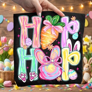 Peut inclure: Sac fourre-tout noir avec les mots "Hip Hop" en lettres color&eacute;es. Le motif comprend une carotte, des oreilles de lapin, des &oelig;ufs de P&acirc;ques et des empreintes de pattes, le tout dans des couleurs pastel vives. Le sac est tenu par une main.