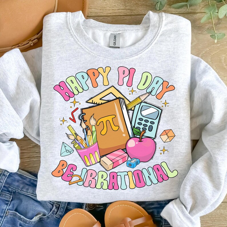 May include: A gray sweatshirt with a colorful graphic design that says "Happy Pi Day Be Irrational". The graphic includes a notebook, pencils, a calculator, a protractor, and an apple.