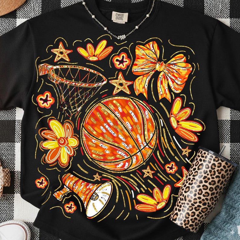 May include: Black t-shirt with a basketball-themed graphic. The design includes a basketball, flowers, stars, a bow, and a megaphone, all in orange, yellow, and gold. A leopard-print tumbler is visible on the right.
