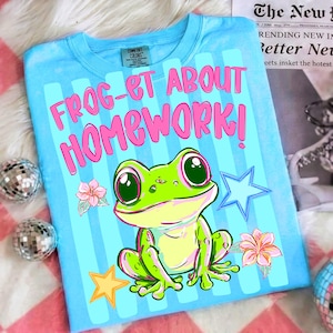 Puede incluir: Camiseta azul claro con el texto rosa "FROG-ET ABOUT HOMEWORK!". La camiseta presenta una rana de dibujos animados, flores y estrellas. La camiseta está hecha de un material suave.
