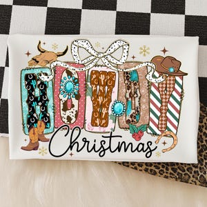 May include: A white rectangular design with the words "HOWDY Christmas" in a Western-themed font. The letters are decorated with cowboy boots, turquoise accents, and a cow skull. A brown cowboy hat and a horseshoe are also included.