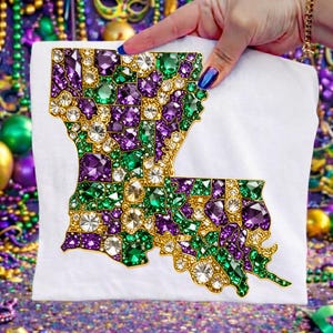 May include: White t-shirt featuring a jeweled outline of the state of Louisiana. The design is filled with purple, green, and clear gemstones, outlined in gold. The shirt is held up by a hand with blue nail polish.