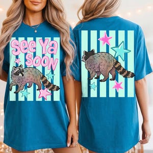 May include: Teal blue t-shirt featuring a raccoon illustration with the text "See Ya Soon". The design includes pink and blue stars and vertical stripes. The raccoon has a striped tail.