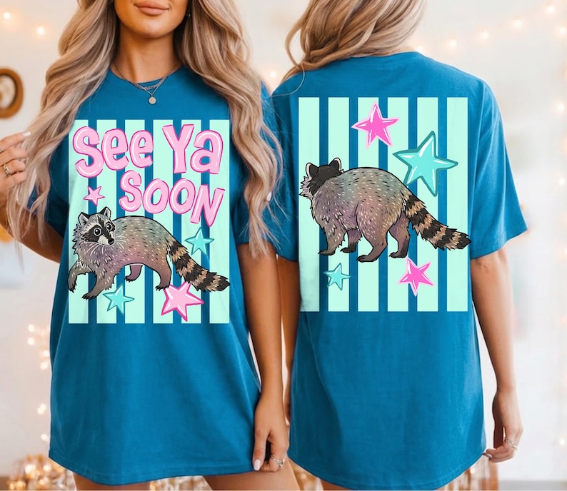 May include: Teal blue t-shirt featuring a raccoon illustration with the text "See Ya Soon". The design includes pink and blue stars and vertical stripes. The raccoon has a striped tail.
