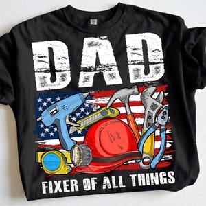 May include: Black t-shirt with a graphic featuring the word "DAD" in white block letters above an American flag design. The design includes tools like a hammer, wrench, and glue gun, with the text "FIXER OF ALL THINGS" below.