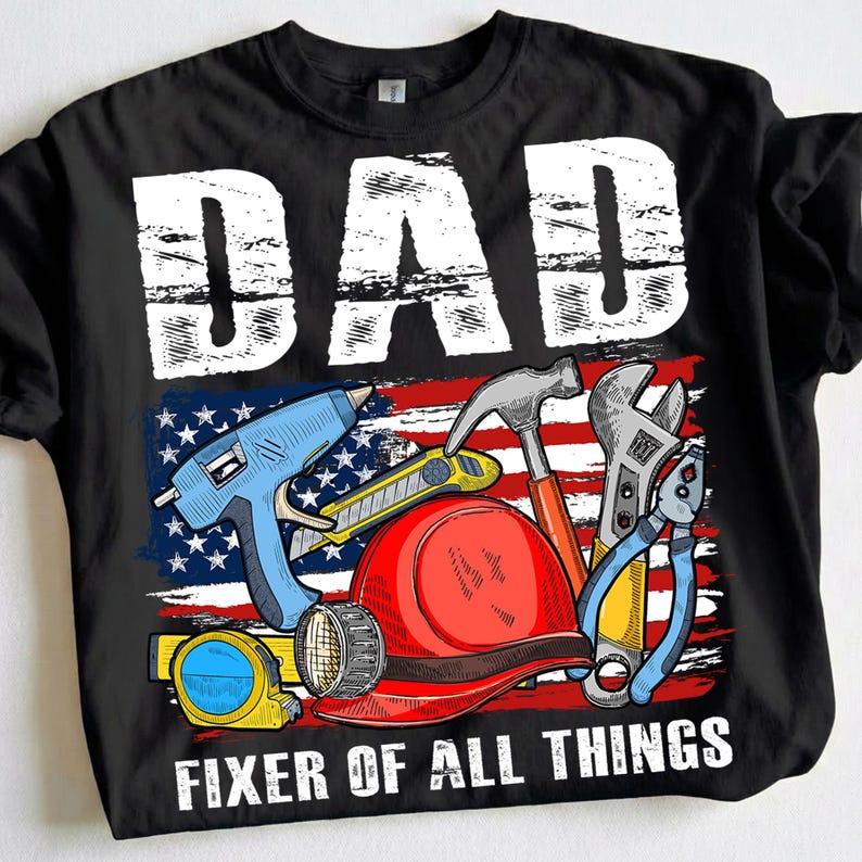 May include: Black t-shirt with a graphic featuring the word "DAD" in white block letters above an American flag design. The design includes tools like a hammer, wrench, and glue gun, with the text "FIXER OF ALL THINGS" below.