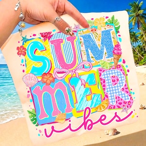 May include: A light beige tote bag with the words "SUMMER vibes" in a colorful, playful design. The letters are outlined in various colors, with a blue striped "U" and a pink bow. The background shows a beach scene.