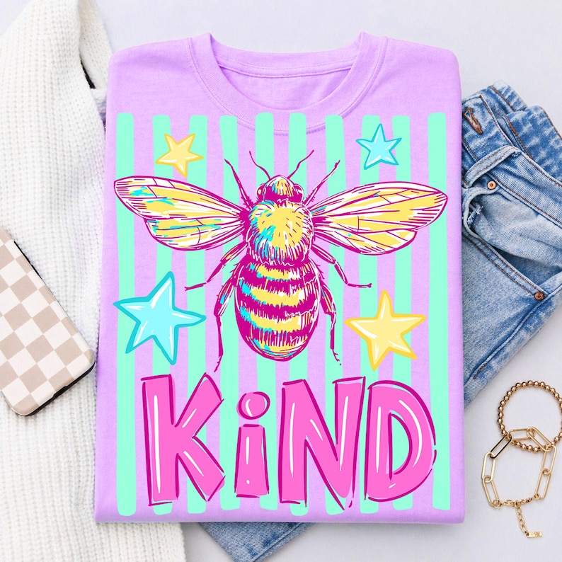 Puede incluir: Una camiseta lila con una ilustraci&oacute;n de abeja colorida, alas amarillas y cuerpo a rayas. El dise&ntilde;o incluye estrellas azules y amarillas y la palabra "KIND" en rosa, sobre un fondo de rayas verdes. Un dise&ntilde;o divertido y brillante.