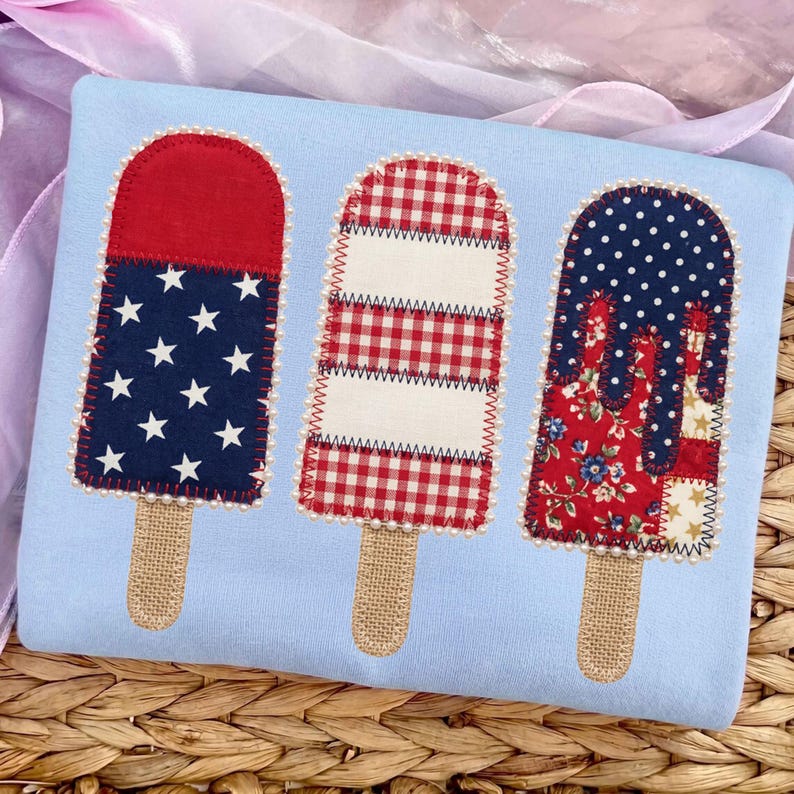 May include: A light blue fabric item with three decorative popsicle designs. Each popsicle has a burlap stick and is decorated with red, white, and blue patriotic patterns, including stars, gingham, and floral prints. The edges are trimmed with small white beads.