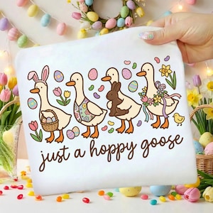 May include: White t-shirt with a whimsical Easter design. Features four cartoon geese, each adorned with Easter-themed accessories like bunny ears, floral patterns, and a chocolate bunny. The text "just a hoppy goose" is written in a playful font.
