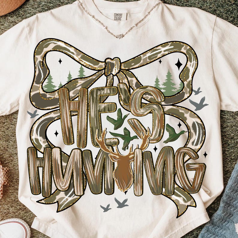 May include: Cream-colored t-shirt with a camouflage bow design and the text "HE'S HUNTING" in a brown, textured font. The design includes a deer silhouette, trees, and flying birds.