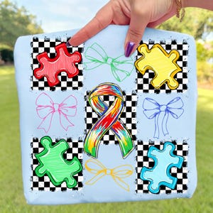 May include: Pale blue tote bag with a design of colourful puzzle pieces, bows, and a ribbon. The design includes red, green, yellow, pink, blue, and orange elements set against a black and white chequered background.