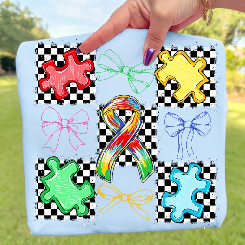 May include: Pale blue tote bag with a design of colourful puzzle pieces, bows, and a ribbon. The design includes red, green, yellow, pink, blue, and orange elements set against a black and white chequered background.