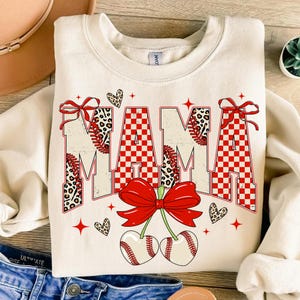 Puede incluir: Sudadera color crema con la palabra "MAMA" en un diseño temático de béisbol. Las letras presentan patrones de cuadros rojos, costuras de béisbol y estampado de leopardo. Los lazos rojos y los gráficos de cerezas de béisbol completan el diseño.