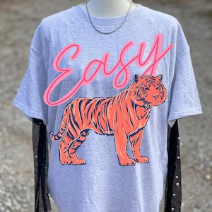 May include: A gray t-shirt featuring a large orange tiger illustration with navy stripes. The word "Easy" is written above the tiger in pink script. The shirt has short sleeves and a crew neck.