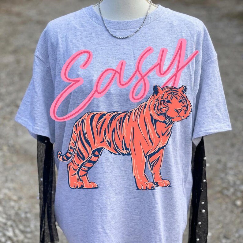 May include: A gray t-shirt featuring a large orange tiger illustration with navy stripes. The word "Easy" is written above the tiger in pink script. The shirt has short sleeves and a crew neck.