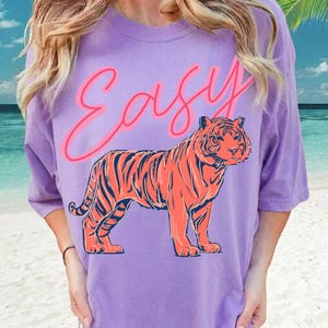 May include: A lilac-colored t-shirt featuring a vibrant orange tiger illustration with navy stripes. Above the tiger, the word "Easy" is written in a cursive, neon pink font. The shirt has short sleeves and a relaxed fit.
